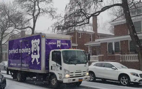 A Perfect Moving truck on a snowy street lined with brick houses, beside a parked white car