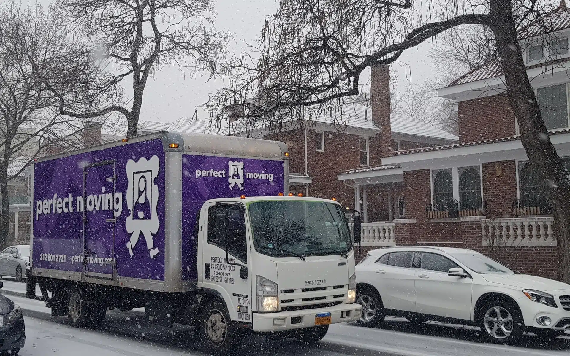 A Perfect Moving truck on a snowy street lined with brick houses, beside a parked white car